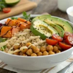 A grain bowl with quinoa, roasted veggies, avocado, chickpeas, and tahini dressing on a wooden table.