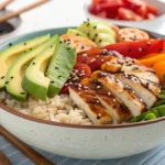 A colorful rice bowl with grilled chicken, avocado, roasted vegetables, sesame seeds, and a drizzle of sauce.