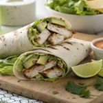 Chicken avocado wrap cut in half, showing grilled chicken, avocado, lettuce, and tomato inside a tortilla, served on a wooden board with a side of dipping sauce.